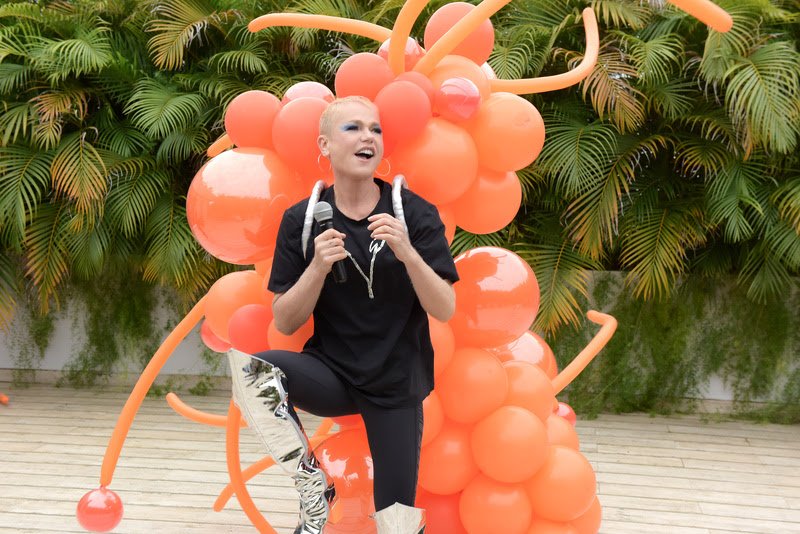 mulher loira de cabelo curto vestida de preto com microfone em frente a arco de bolas laranja para divulgar a apresentação da artista Xuxa no Rock in Rio