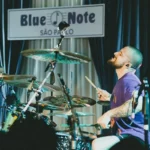 Baterista Eloy Casagrande no espaço Blue Note, em São Paulo, tocando bateria. Eloy está na segunda metade da tela, rosto inclinado para cima, enquadrado de perfil.