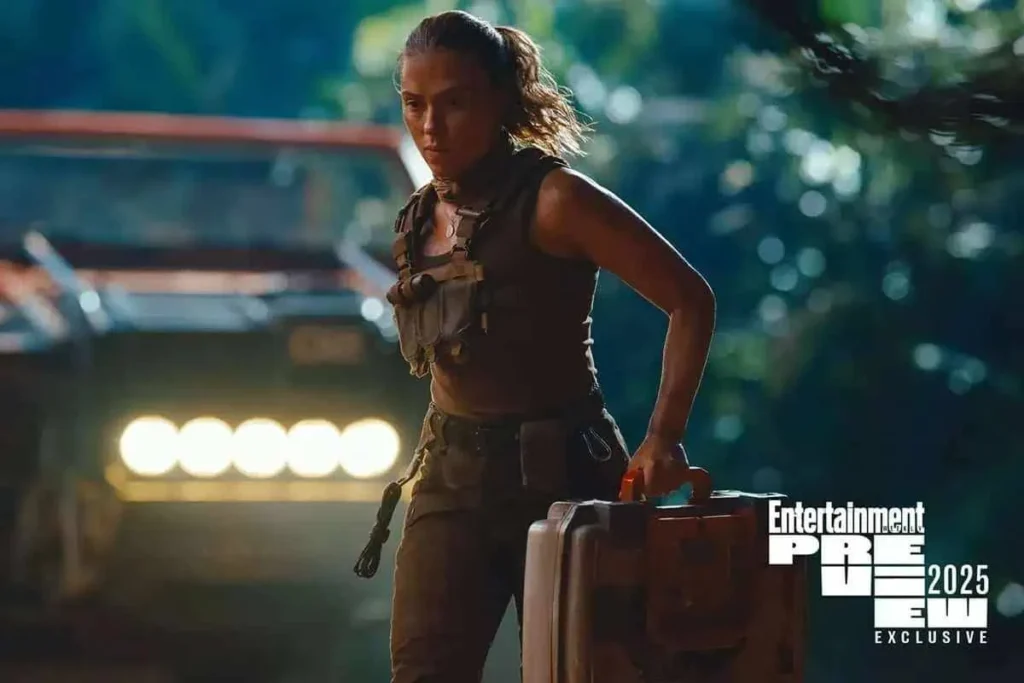 Jurassic World Rebirth | Elenco Passou por Perigos durante as Filmagens Sem Saber 3 Personagem de Jurassic World Rebirth em um matagal, trajada com roupas de exploração, carregando uma mala. Há um carro atrás.