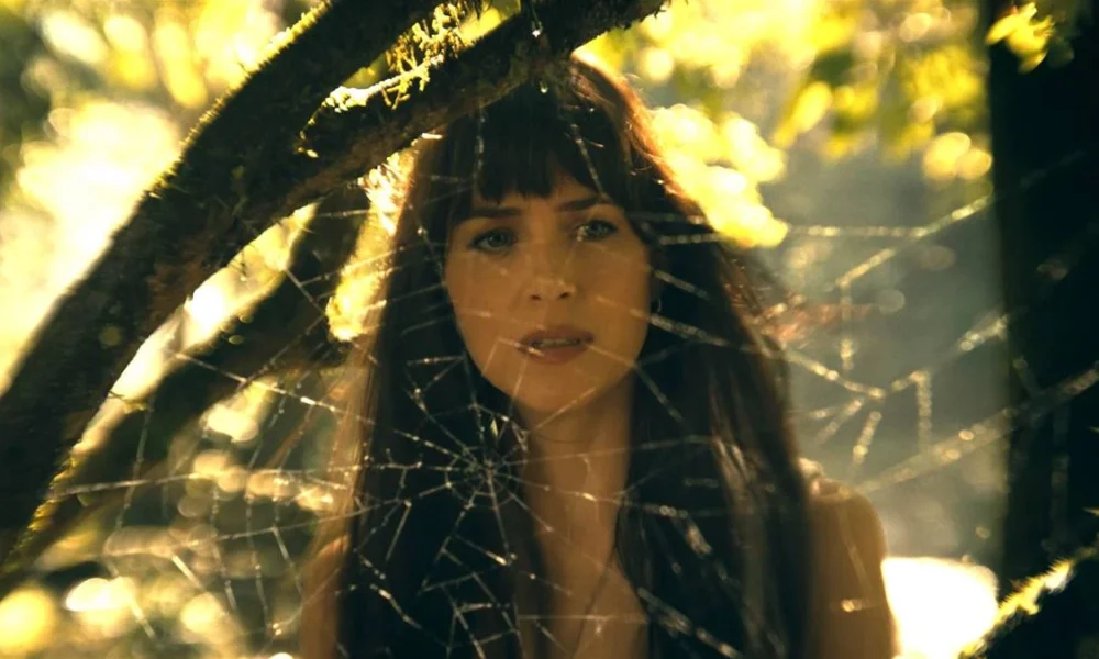 A protagonista de Madame Teia, conforme vista suavemente embaçada por detrás de uma teia de aranha entre árvores em um dia de sol.