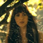 A protagonista de Madame Teia, conforme vista suavemente embaçada por detrás de uma teia de aranha entre árvores em um dia de sol.