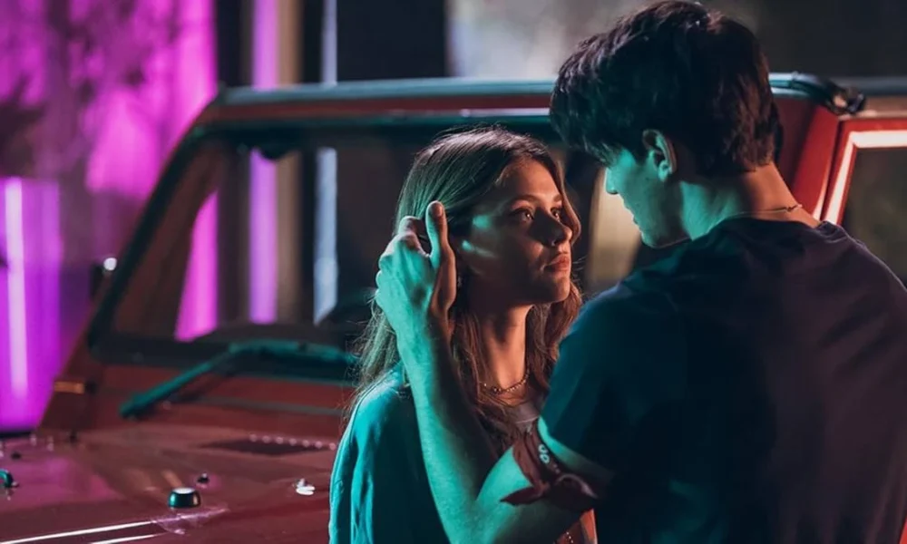 Captura de tela promocional dos protagonistas de "Sua Culpa" no filme anterior: "Minha Culpa", pela Prime Video. Imagem com luzes neon e os protagonistas se olhando, e acariciando, em frente a um carro.