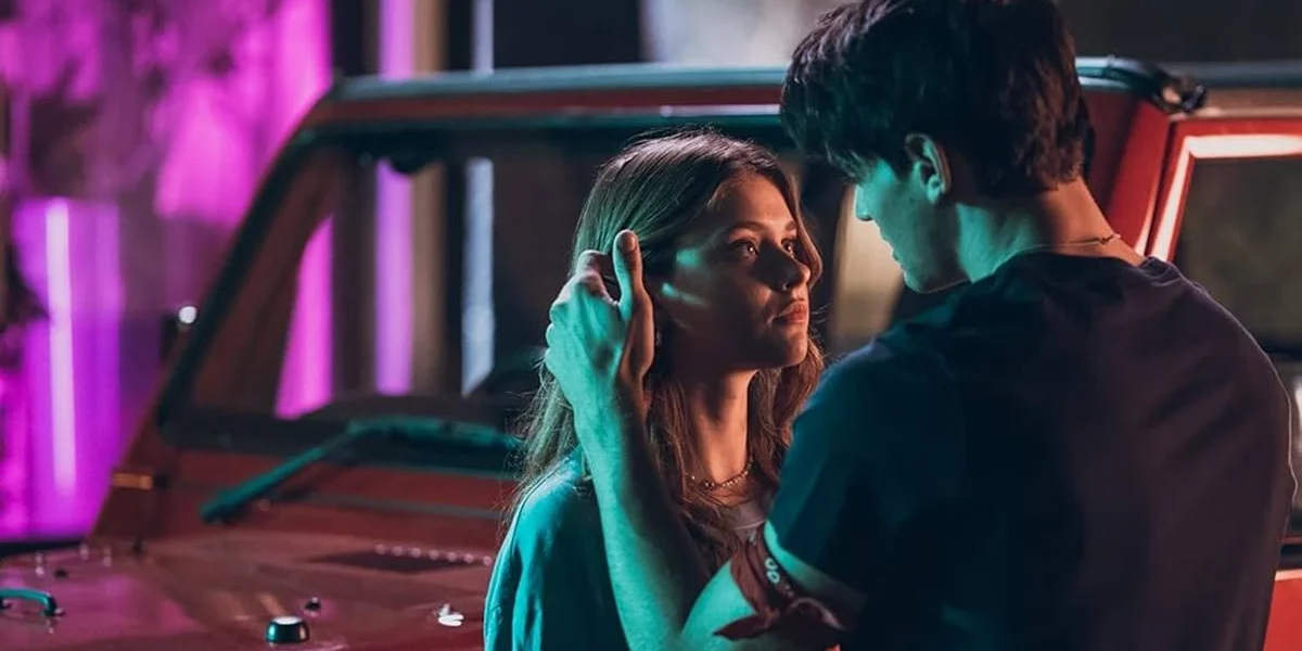 Captura de tela promocional dos protagonistas de "Sua Culpa" no filme anterior: "Minha Culpa", pela Prime Video. Imagem com luzes neon e os protagonistas se olhando, e acariciando, em frente a um carro.