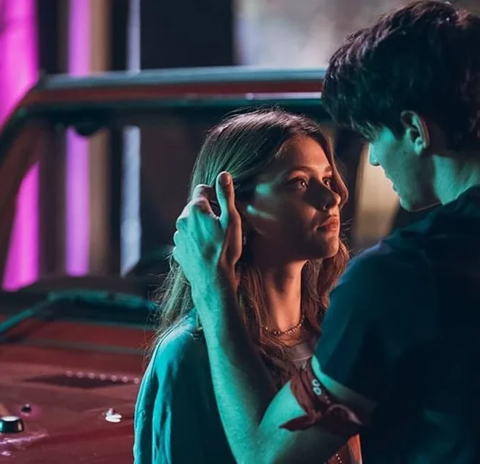 Captura de tela promocional dos protagonistas de "Sua Culpa" no filme anterior: "Minha Culpa", pela Prime Video. Imagem com luzes neon e os protagonistas se olhando, e acariciando, em frente a um carro.