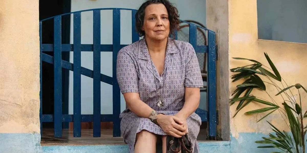 A personagem Moema (Ana Beatriz Nogueira) na novela "Mania de Você", da TV Globo. Ela está sentada no portão de uma casa, com as mãos sobre o colo.