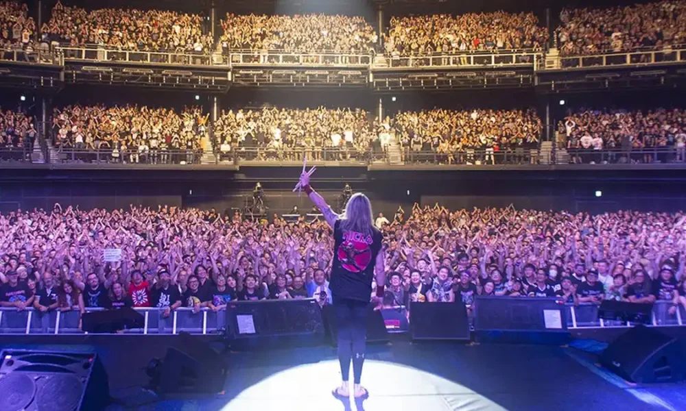 Fotografia tirada por trás do palco, com o baterista Nicko McBrain agradecendo, de braço esquerdo levantado e baquetas erguidas, aos fãs do Iron Maiden.