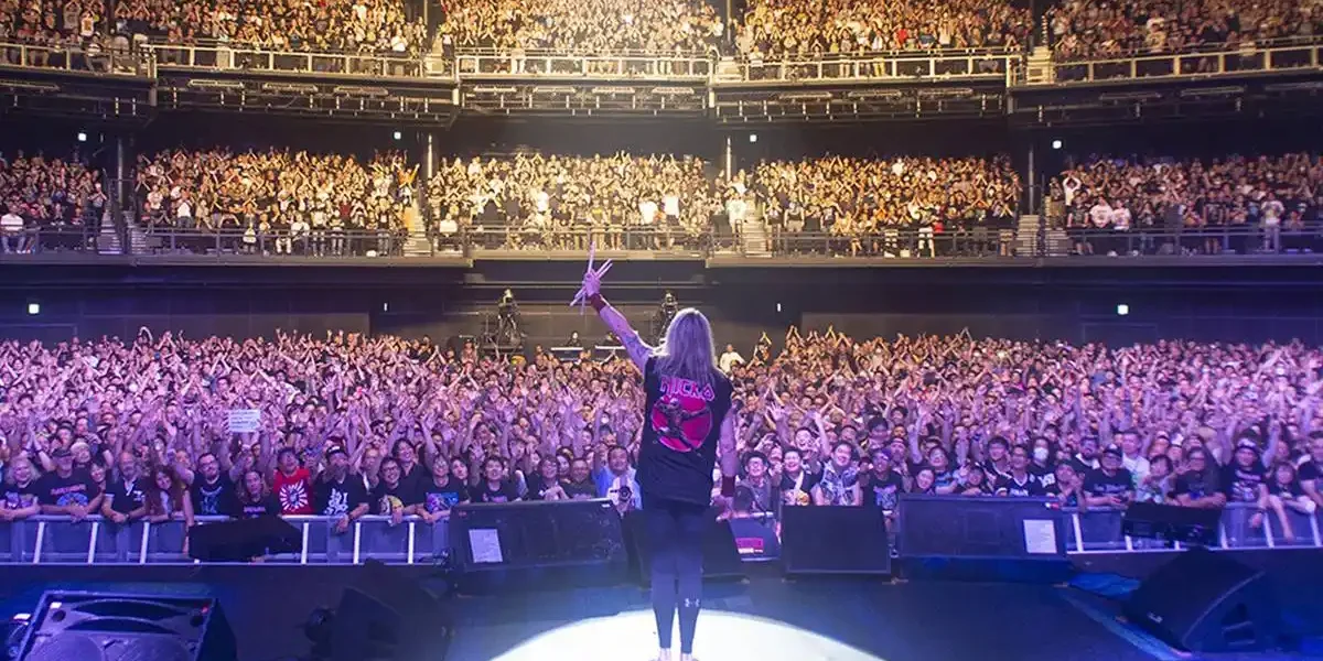 Fotografia tirada por trás do palco, com o baterista Nicko McBrain agradecendo, de braço esquerdo levantado e baquetas erguidas, aos fãs do Iron Maiden.