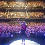 Fotografia tirada por trás do palco, com o baterista Nicko McBrain agradecendo, de braço esquerdo levantado e baquetas erguidas, aos fãs do Iron Maiden.