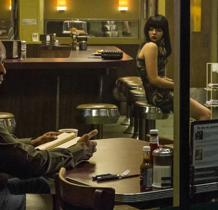 Denzel Washington como o protagonista da trilogia "O Protetor", em seu primeiro filme, de 2014, sentado em um bar lendo.