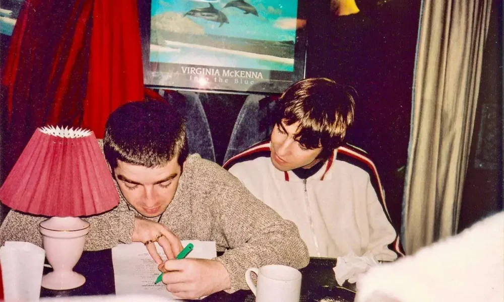 Fotografia com os irmãos Gallagher, da banda Oasis, em uma mesa. Um olha o outro escrever algo com uma caneta verde.