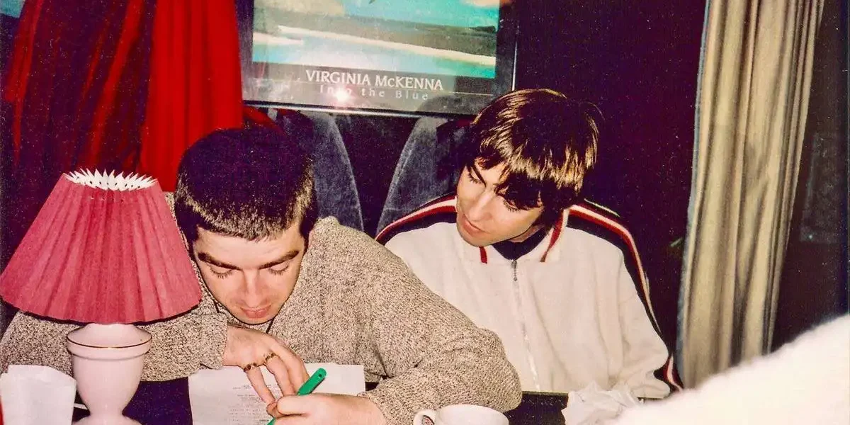 Fotografia com os irmãos Gallagher, da banda Oasis, em uma mesa. Um olha o outro escrever algo com uma caneta verde.