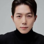 Fotografia do ator Park Min-jae, do busto para cima, olhando para a câmera. Park está com um sorriso sério e vestido de preto.