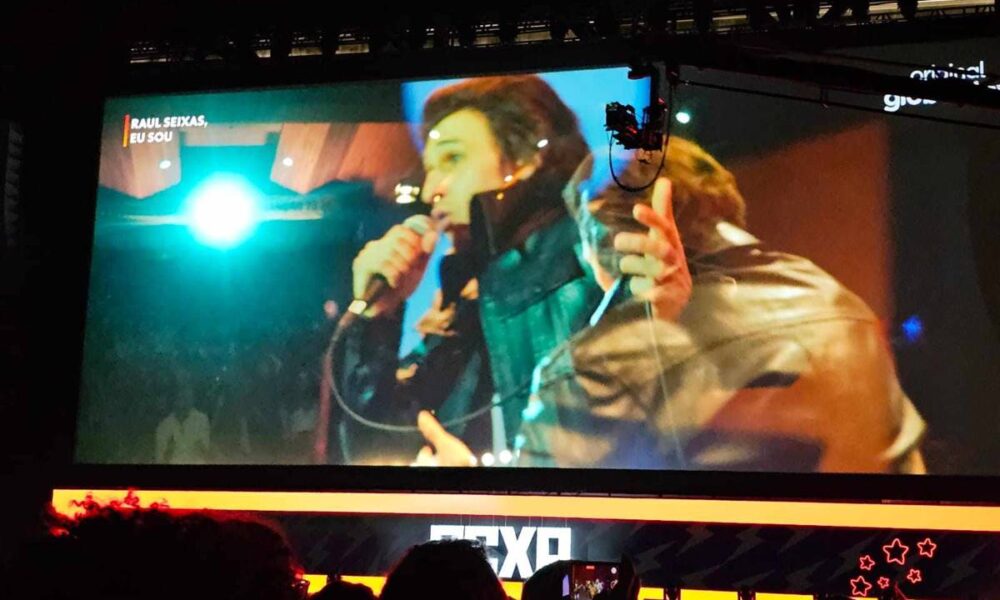 Fotografia do painel de "Raul Seixas, Eu Sou", com trailer sendo exibido pela Globoplay. Um dos atores está caracterizado e cantando no papel do protagonista.