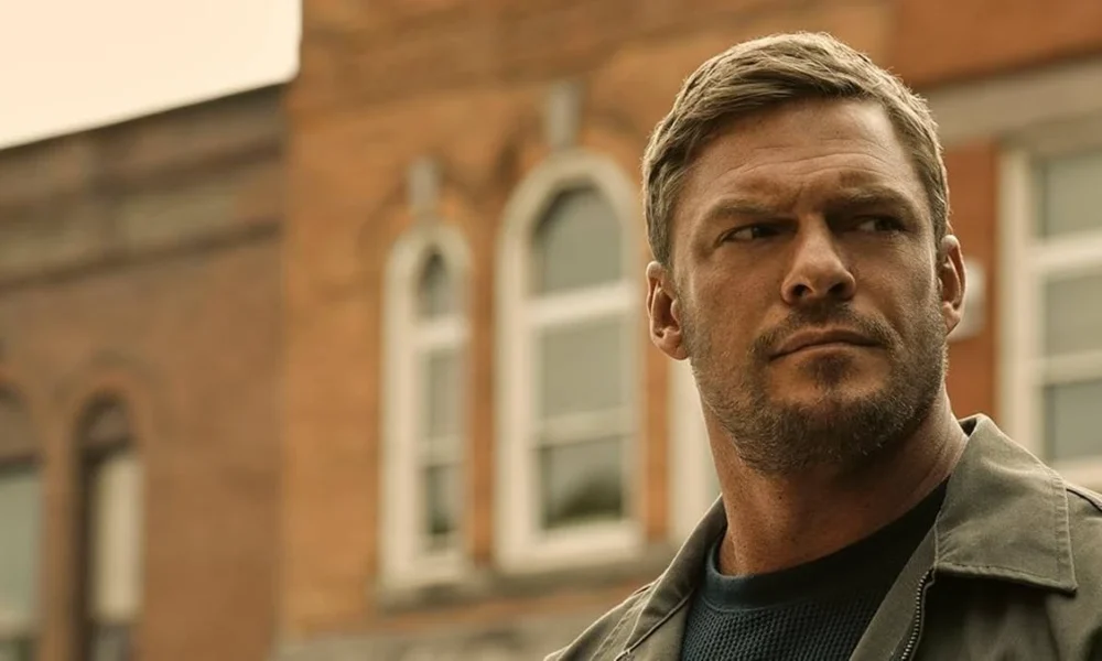 Protagonista da série Reacher em cena da segunda temporada, com ele na segunda metade do enquadramento e o fundo, um prédio de tijolos com janelas largas, e o céu nublado. Feição séria.