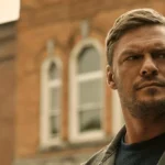 Protagonista da série Reacher em cena da segunda temporada, com ele na segunda metade do enquadramento e o fundo, um prédio de tijolos com janelas largas, e o céu nublado. Feição séria.