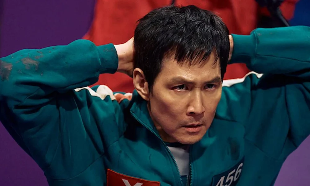 Gi-hun durante segunda temporada de Round 6 ("The Squid Game") com as mãos atrás da cabeça, postura de rendido, e uniformizado para o jogo.