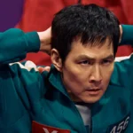 Gi-hun durante segunda temporada de Round 6 ("The Squid Game") com as mãos atrás da cabeça, postura de rendido, e uniformizado para o jogo.