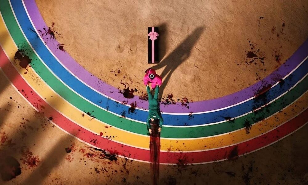 Uma pista arco-íris ensanguentada para a segunda temporada de Round 6. Um dos minions de vermelho puxa um cadáver para um caixão em forma de caixa de presente.