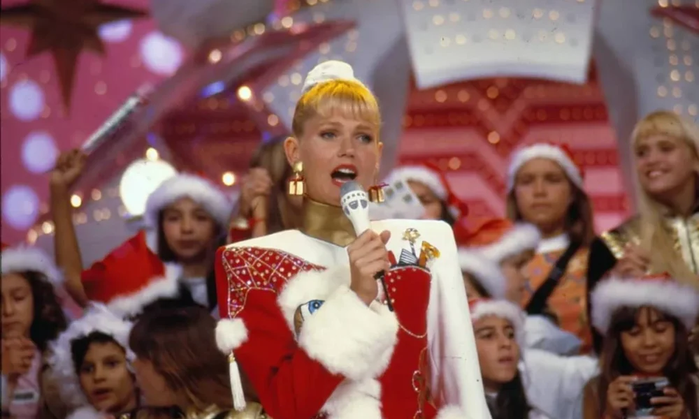 Imagem da cantora e apresentadora Xuxa em um especial de natal do "Xou da Xuxa". Xuxa está vestida com roupas natalinas vermelhas e brancas. A plateia, atrás, está com gorros vermelhos de natal.