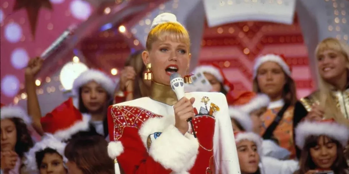 Imagem da cantora e apresentadora Xuxa em um especial de natal do "Xou da Xuxa". Xuxa está vestida com roupas natalinas vermelhas e brancas. A plateia, atrás, está com gorros vermelhos de natal.