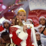 Imagem da cantora e apresentadora Xuxa em um especial de natal do "Xou da Xuxa". Xuxa está vestida com roupas natalinas vermelhas e brancas. A plateia, atrás, está com gorros vermelhos de natal.