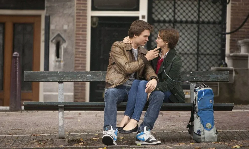 Personagens de "A Culpa é das Estrelas", filme de 2014: Augustus Waters (Ansel Elgort) e Hazel Grace Lancaster (Shailene Woodley), respectivamente à esquerda e direita. Ela está com aparelhos para ajudarem-na a respirar e ele segura o queixo dela de maneira carinhosa. Ambos estão sentados em um banco ao ar livre.