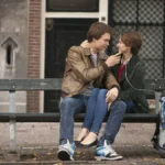 Personagens de "A Culpa é das Estrelas", filme de 2014: Augustus Waters (Ansel Elgort) e Hazel Grace Lancaster (Shailene Woodley), respectivamente à esquerda e direita. Ela está com aparelhos para ajudarem-na a respirar e ele segura o queixo dela de maneira carinhosa. Ambos estão sentados em um banco ao ar livre.