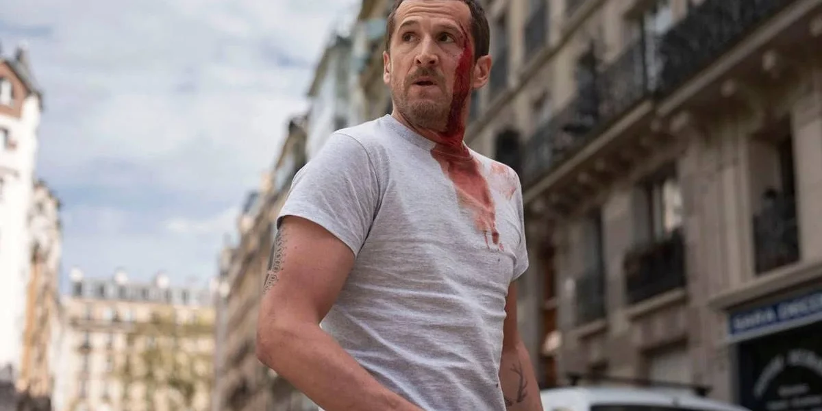 Protagonista de "Ad Vitam" (2025) ensanguentado, na metade esquerda do corpo, em uma rua. Ele olha para longe e está na frente de uma rua com arquitetura clássica francesa. Está de dia e ele veste branco.