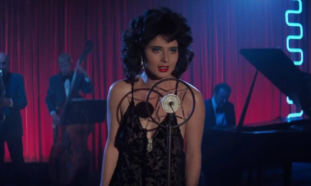 Isabella Rossellini como Dorothy Vallens, protagonista de Veludo Azul, performando uma música. Há pianistas atrás e um microfone em sua frente.