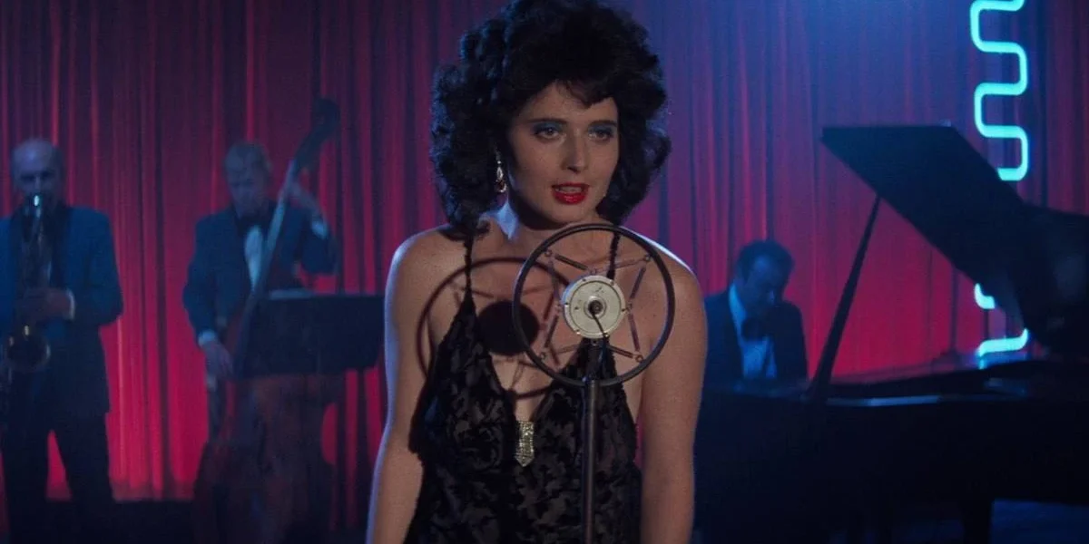 David Lynch | As Obras Mais Marcantes: 9 Filmes Essenciais Isabella Rossellini como Dorothy Vallens, protagonista de Veludo Azul, performando uma música. Há pianistas atrás e um microfone em sua frente.