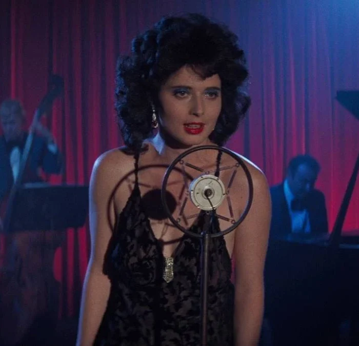 Isabella Rossellini como Dorothy Vallens, protagonista de Veludo Azul, performando uma música. Há pianistas atrás e um microfone em sua frente.