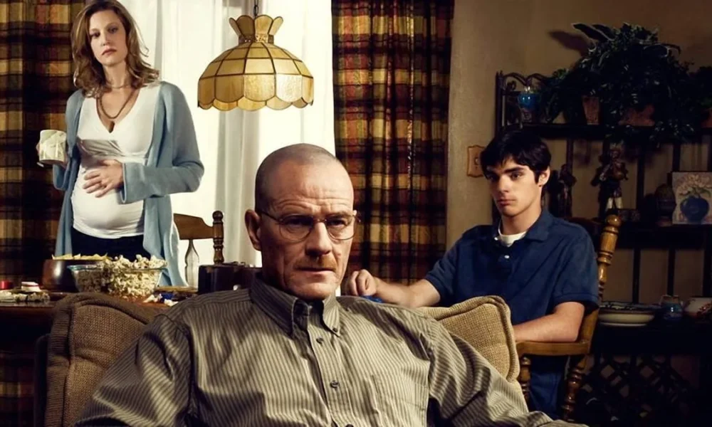 Walter White, da série The Breaking Bad, sentado em uma poltrona na sua casa. Atrás dele, esposa e filho tomam café e o olham inquietos.