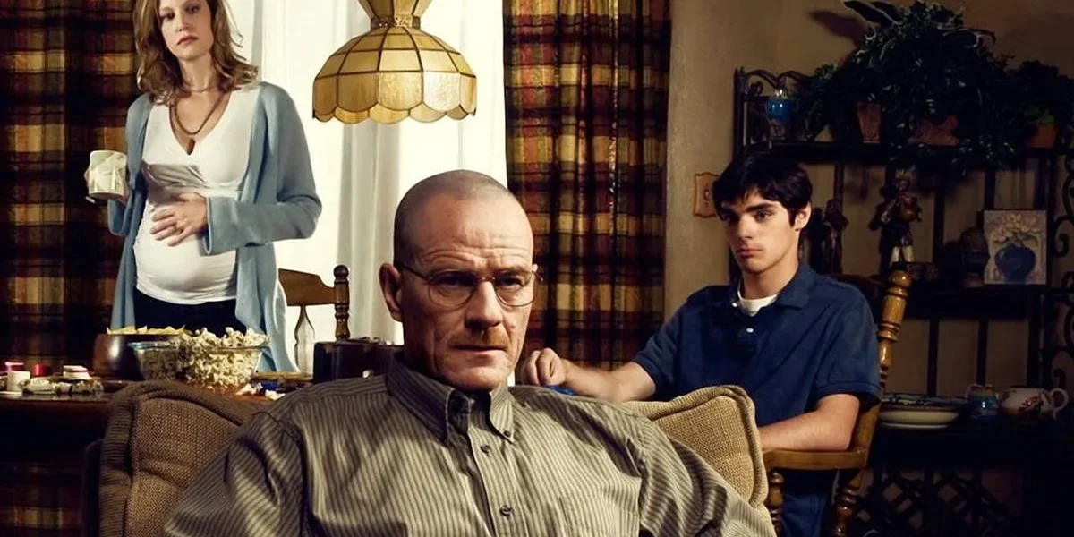 Walter White, da série The Breaking Bad, sentado em uma poltrona na sua casa. Atrás dele, esposa e filho tomam café e o olham inquietos.