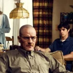 Walter White, da série The Breaking Bad, sentado em uma poltrona na sua casa. Atrás dele, esposa e filho tomam café e o olham inquietos.