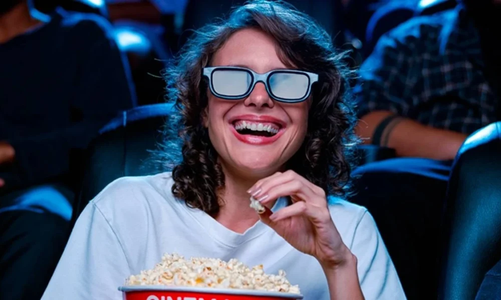 Uma mulher de cabelos ondulados e vestida de branco com óculos 3D, comendo pipoca com a mão esquerda, rindo, assistindo um cinema em uma sala Cinemark.