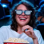 Uma mulher de cabelos ondulados e vestida de branco com óculos 3D, comendo pipoca com a mão esquerda, rindo, assistindo um cinema em uma sala Cinemark.