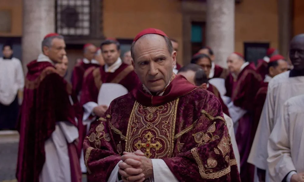 Ralph Fiennes como protagonista Lawrence, em destaque em patio cercado de cardeais no filme "Conclave" (2024). Personagem está com dedos juntos.