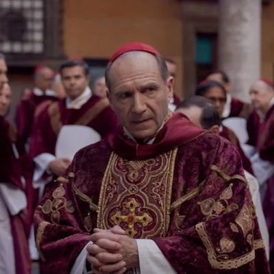 Ralph Fiennes como protagonista Lawrence, em destaque em patio cercado de cardeais no filme "Conclave" (2024). Personagem está com dedos juntos.