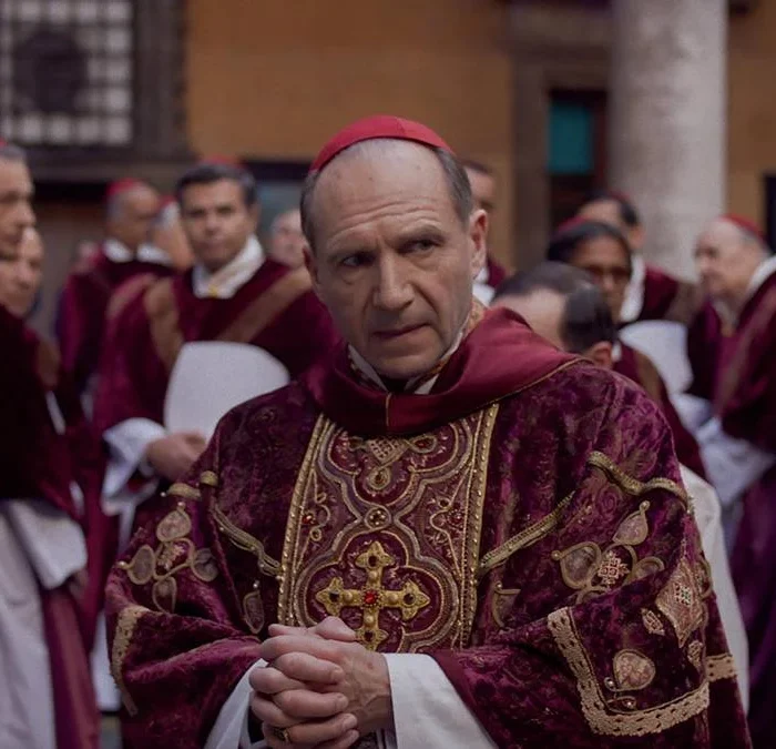 Ralph Fiennes como protagonista Lawrence, em destaque em patio cercado de cardeais no filme "Conclave" (2024). Personagem está com dedos juntos.