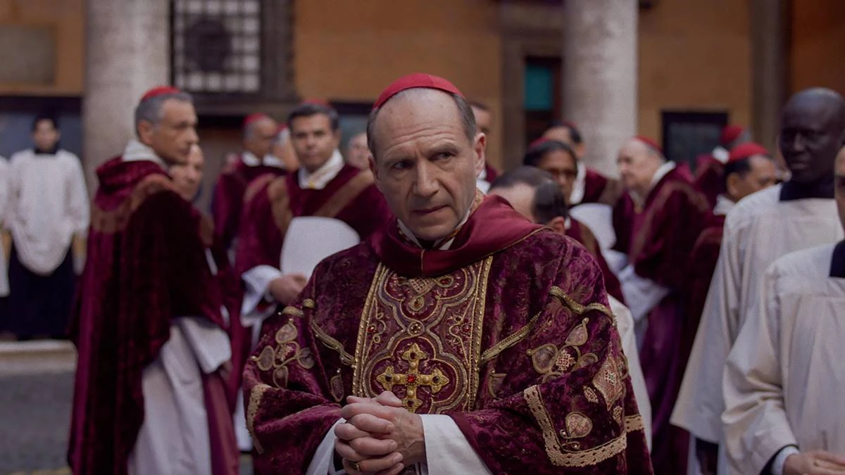 Conclave | Dúvidas, Segredos e Disputa Pelo Poder em Meio à Fé 2 Ralph Fiennes como protagonista Lawrence, em destaque em patio cercado de cardeais no filme "Conclave" (2024). Personagem está com dedos juntos.
