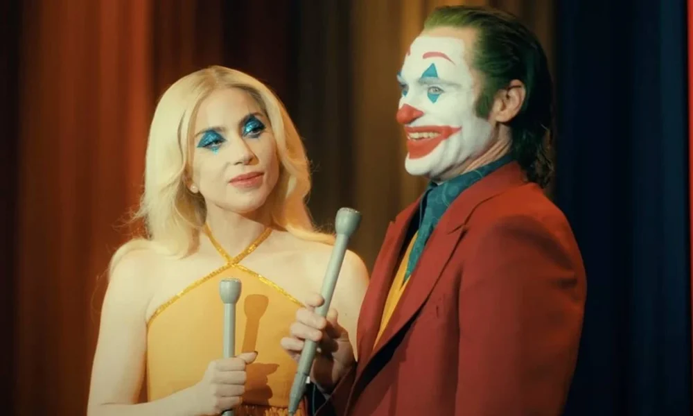 Lady Gaga e Joaquin Phoenix no filme "Coringa: Delírio a 2" (Joker 2) como os personagens Arlequina e Coringa. Ambos estão com maquiagem carregada e vestidos para um programa de televisão.