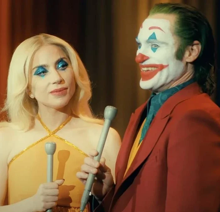 Lady Gaga e Joaquin Phoenix no filme "Coringa: Delírio a 2" (Joker 2) como os personagens Arlequina e Coringa. Ambos estão com maquiagem carregada e vestidos para um programa de televisão.