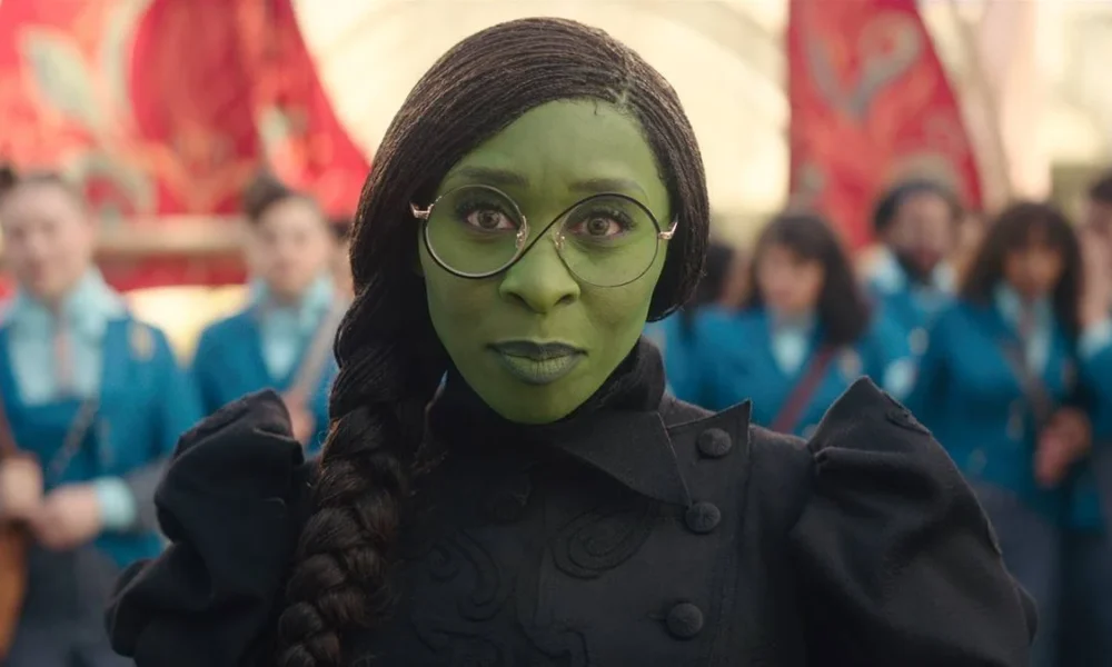 Cynthia Erivo como a protagonista Elphaba de "The Wicked", filme um. A personagem de pele verde está com seus óculos de lentes redondas e com um sorriso discreto à câmera.