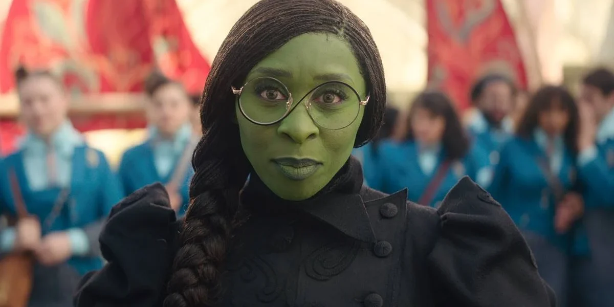 Cynthia Erivo como a protagonista Elphaba de "The Wicked", filme um. A personagem de pele verde está com seus óculos de lentes redondas e com um sorriso discreto à câmera.