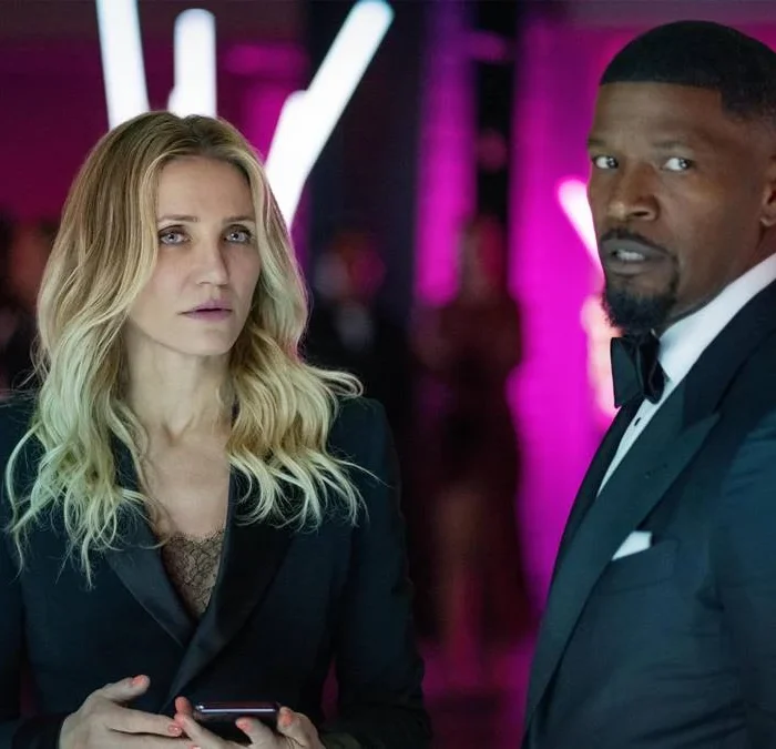 Cameron Diaz e Jamie Foxx, respectivamente à esquerda e direita, vestidos de trajes sociais, em um ambiente de fundo neon roxo, interpretando seus papéis em "De Volta À Ação" (2025).