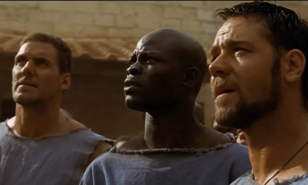 Ator DJimon Hounsou como visto em cena de "Gladiador" (2000), onde ele está ao centro, olhando longe da câmera, à esquerda da tela.