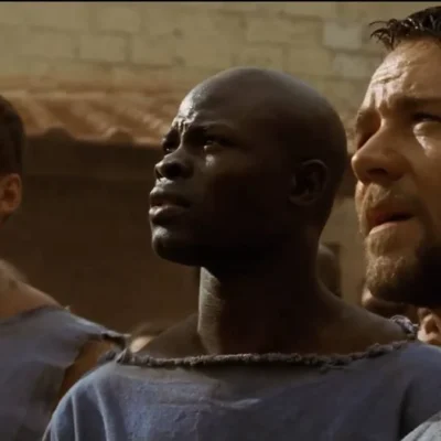 Ator DJimon Hounsou como visto em cena de "Gladiador" (2000), onde ele está ao centro, olhando longe da câmera, à esquerda da tela.