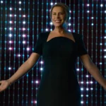 Karla Sofía Gascón como a protagonista de "Emilia Pérez", com um vestido preto com manga curta, um colar de ouro discreto, e sorrindo e braços abertos. Atrás dela, luzes em forma de cortina.