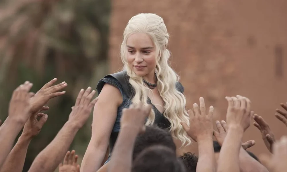 Daenerys Targaryen, com roupa azul e cabelo solto, cercada por seguidores querendo tocar nela, em "Game of Thrones".