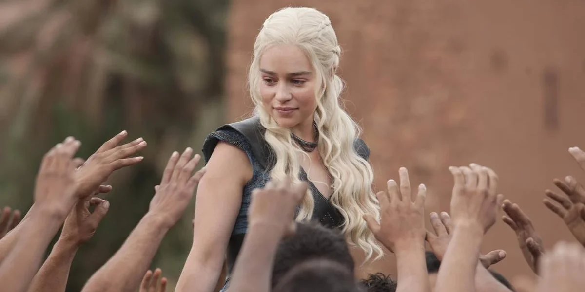 Daenerys Targaryen, com roupa azul e cabelo solto, cercada por seguidores querendo tocar nela, em "Game of Thrones".